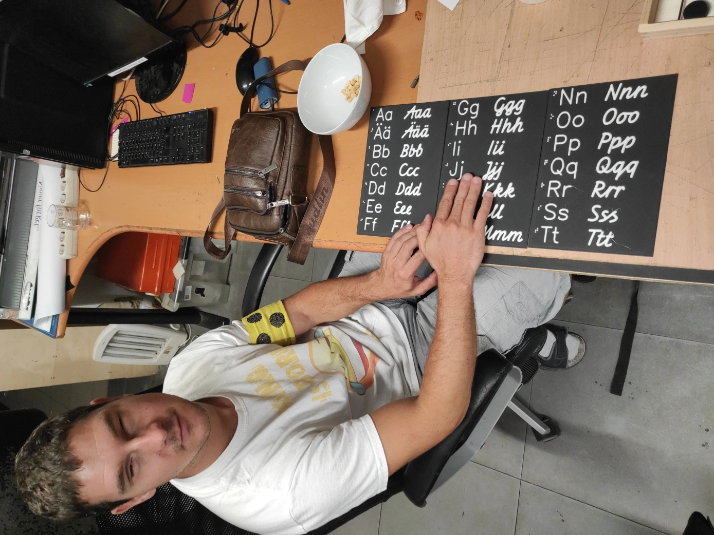 Eine Person hat beide Hände auf schwarzen Tafeln. Auf den Tafeln steht das Alphabet in Braille, Großbuchstaben, Kleinbuchstaben in Druckbuchstaben und in Schreibschrift. Die Tafeln liegen auf einem Tisch gemeinsam mit einer Tasche, einer Schüssel mit Nüssen und Computerausstattung. Die Person trägt eine gelbe Armschleife mit drei schwarzen Punkten.