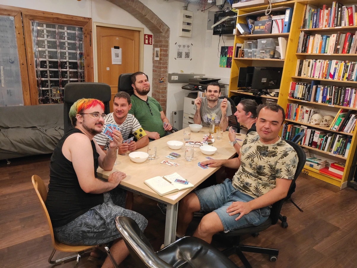 Sechs Personen sitzen an einem Schreibtisch, halten Karten in den Händen und lächeln zum größten Teil in die Kamera. Sie tragen T-Shirts und sitzen auf Bürostühlen. Auf dem Tisch stehen Schüsseln, Gläser, Süßigkeiten und Karten. Eine Person trägt eine Armbinde mit dem Verkehrsschutzzeichen für Menschen mit Blindheit oder Sehbehinderung in Österreich, drei im Dreieck angeordnete schwarze Punkte auf gelbem Grund. Eine andere Person hat eine Frisur in Regenbogenfarben. Im Hintergrund stehen ein Sofa, Elektronik und Bücher.