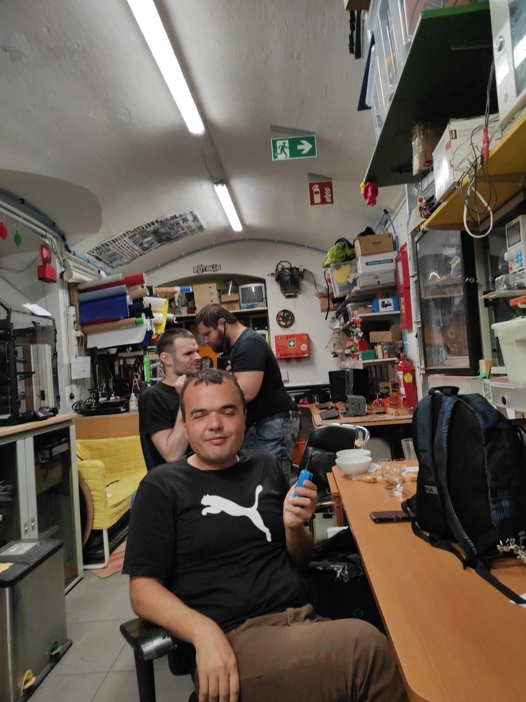 Drei junge Männer in schwarzen T-Shirts in einer Werkstatt. Im Vordergrund sitzt einer der Männer und hält ein Funkgerät in Richtung Kamera. Ein Mann sitzt hinter dem Mann im Vordergrund. Sein Funkgerät wird zur hälfte vom Mann im Vordergrund verdeckt. Dahinter steht ein dritter Mann mit Bart. Der Raum ist länglich und wird von Neonröhren an der gewölbten Decke beleuchtet. Die Wände sind weiß gestrichen. An den Wänden stehen Tische, Schränke und ein gelbes Sofa. Über den Tischen befinden sich vollgeräumte Regalfächer. Ein Fluchtwegschild, Feuerlöscher und Erste Hilfe Koffer sind erkennbar.